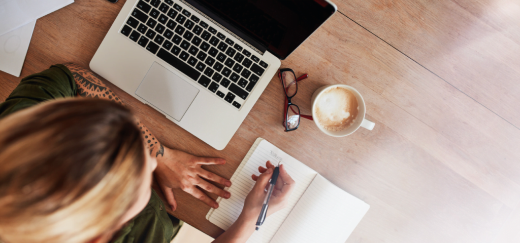 woman writing on desk with a cup of coffee, open laptop and eyeglasses